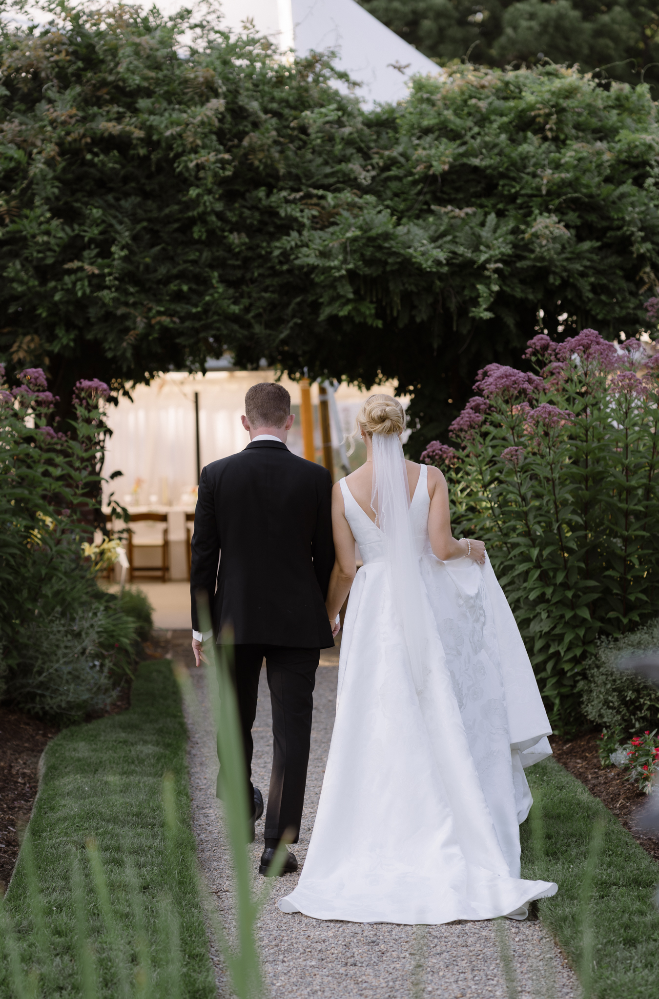 Bride and groom walking hand in hand through a garden toward their wedding reception tent