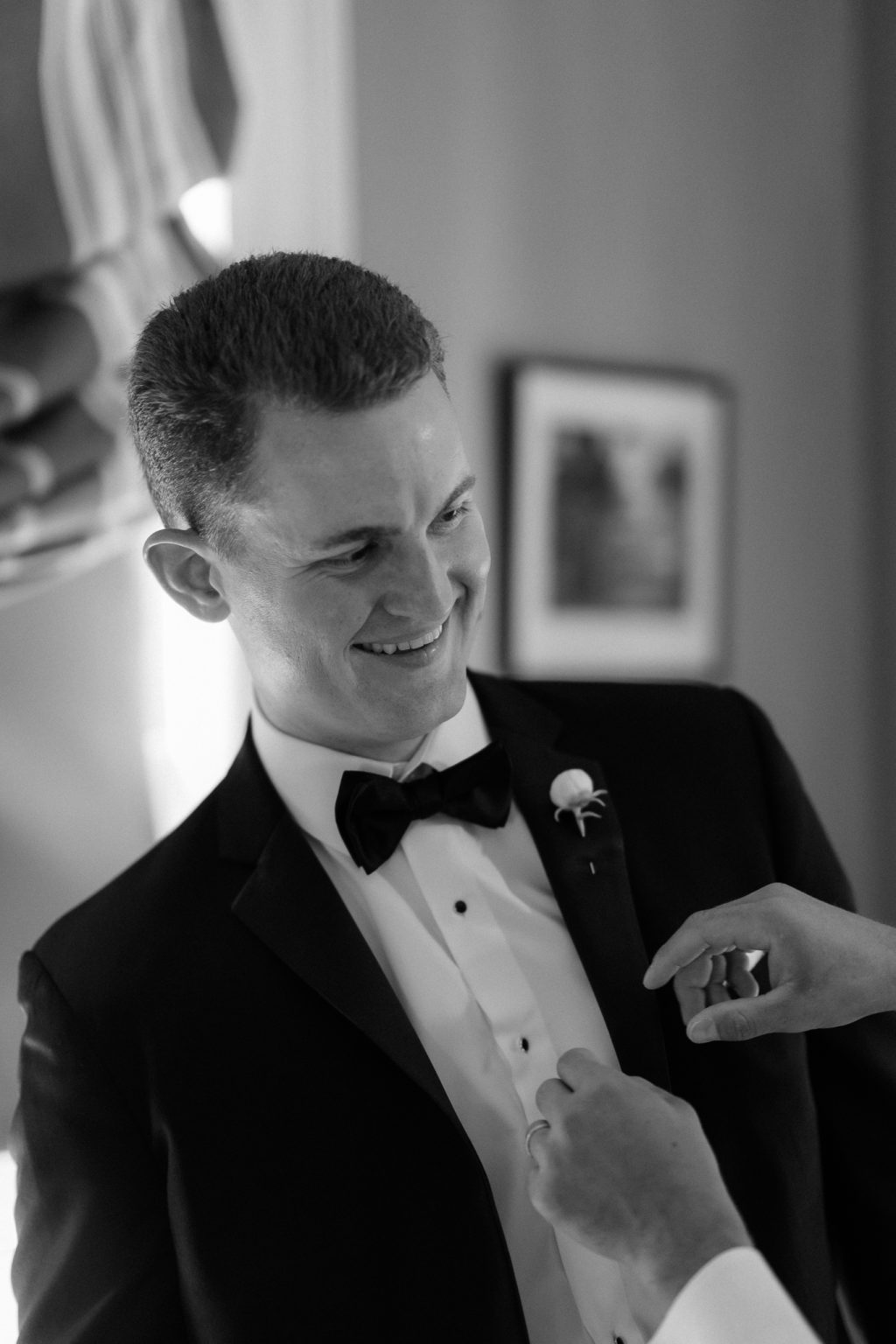 Groom smiling as his bow tie is adjusted while getting ready, black and white photo