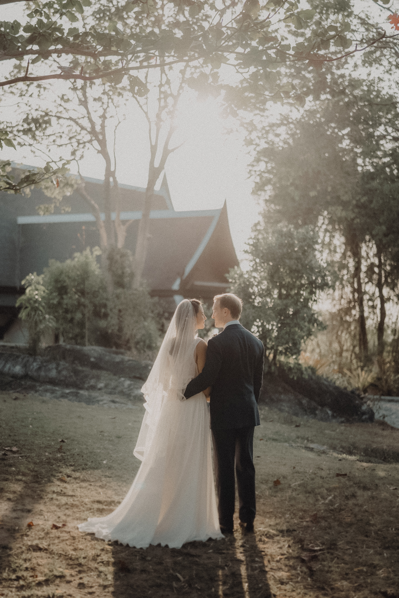 Bride and groom standing together in golden backlight beside a traditional pavilion