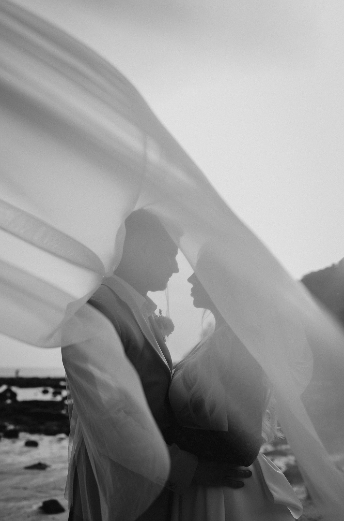 Creative black & white portrait of the wedding couple under the brides veil as it blows in the wind